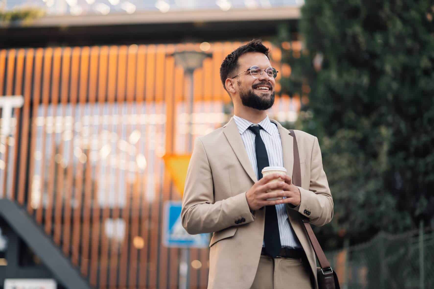 A smiling man in a tan suit holds a coffee cup while standing outdoors near a modern building.