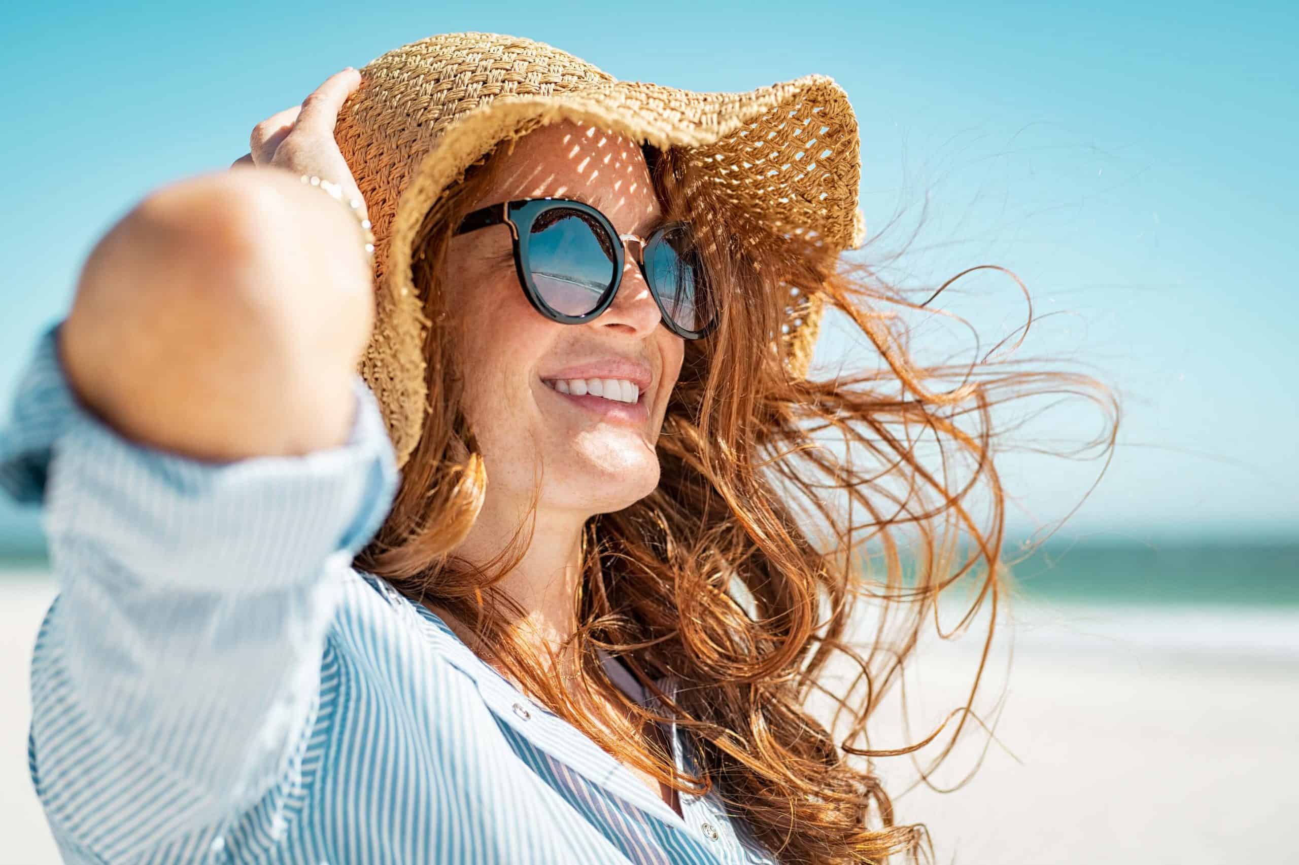 A smiling woman in sunglasses and a straw hat holds her hat in the wind on a sunny Marina Del Rey beach.